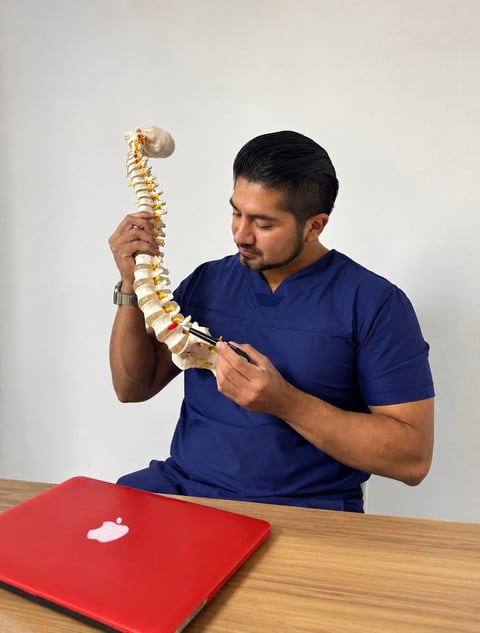 Healthcare professional in blue scrubs examining a spinal column model at a desk with a red laptop